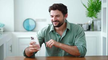 Charming young man, in casual wear using smartphone for a communication by link, smiling to his interlocutor, having fun dialogue, looking at web camera while relaxing in the cozy home interior video