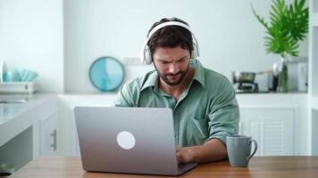 Young bearded Caucasian man wearing wireless headphones, is relaxing, listening to the music while working on a new project on laptop computer from home. Portrait of freelancer in home office video