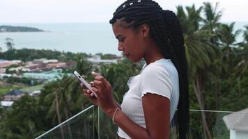 Relaxed multi-ethnic woman using mobile phone, typing text on her smartphone, standing on a balcony with an enchanting view of tropical Asian nature. Online, internet, distance communication concept video