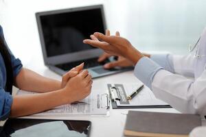 Doctor and patient sitting near each other at the wooden desk in clinic. Female physician's pointing to a records form. Medicine concept photo