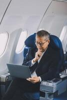 Attractive Asian male passenger of airplane sitting in comfortable seat while working laptop and tablet with mock up area using wireless connection. Travel in style, work with grace. photo
