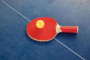 Red table tennis racket with a yellow ball between them on a blue background, ping pong sports equipment, symmetry concept regarding the center of the table with a white line, top view, photo