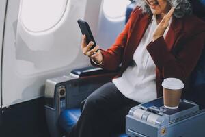 Portrait of a successful Asian businesswoman or entrepreneur in a formal suit on an airplane sitting in business class using a phone, computer laptop. Travel in style, work with grace. photo