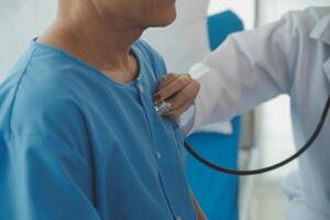 Doctor using stethoscope to listen senior man heart during examination in hospital room and wearing visor as safety precaution against coronavirus. Medical control for infections, disease. photo