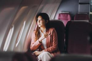 Portrait of a successful Asian businesswoman or entrepreneur in a formal suit on an airplane sitting in business class using a phone, computer laptop. Travel in style, work with grace. photo
