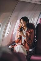 Portrait of a successful Asian businesswoman or entrepreneur in a formal suit on an airplane sitting in business class using a phone, computer laptop. Travel in style, work with grace. photo