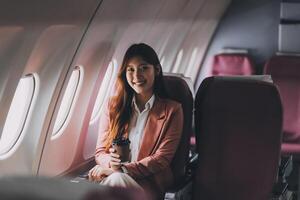 Portrait of a successful Asian businesswoman or entrepreneur in a formal suit on an airplane sitting in business class using a phone, computer laptop. Travel in style, work with grace. photo