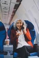 Portrait of a successful Asian businesswoman or entrepreneur in a formal suit on an airplane sitting in business class using a phone, computer laptop. Travel in style, work with grace. photo