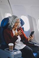 Portrait of a successful Asian businesswoman or entrepreneur in a formal suit on an airplane sitting in business class using a phone, computer laptop. Travel in style, work with grace. photo