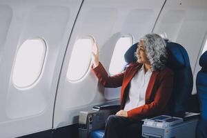 Portrait of a successful Asian businesswoman or entrepreneur in a formal suit on an airplane sitting in business class using a phone, computer laptop. Travel in style, work with grace. photo