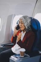 Portrait of a successful Asian businesswoman or entrepreneur in a formal suit on an airplane sitting in business class using a phone, computer laptop. Travel in style, work with grace. photo