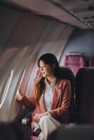 Portrait of a successful Asian businesswoman or entrepreneur in a formal suit on an airplane sitting in business class using a phone, computer laptop. Travel in style, work with grace. photo