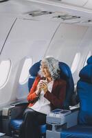 Portrait of a successful Asian businesswoman or entrepreneur in a formal suit on an airplane sitting in business class using a phone, computer laptop. Travel in style, work with grace. photo