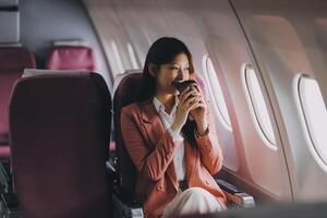 Portrait of a successful Asian businesswoman or entrepreneur in a formal suit on an airplane sitting in business class using a phone, computer laptop. Travel in style, work with grace. photo