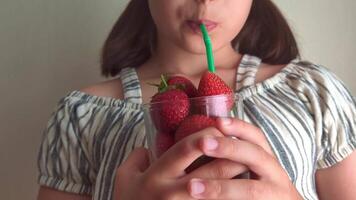 A cute little girl holding a glass of strawberries in both hands and drinking juice through a straw. video