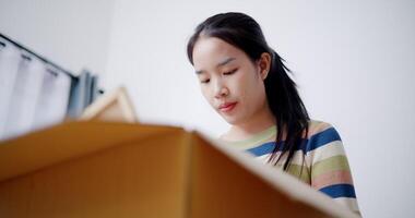 Handheld low angle view shot, Young Asian woman is taking notes while checking belongings in cardboard boxes in preparation for moving to a new house. Moving house and lifestyle concept. video