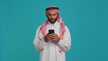 Islamic man works on his mobile gadget, surfing through social media platforms or online pages. Middle eastern guy in customary apparel uses smartphone to access global web, feeling excited. photo