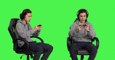 Asian young man plays mobile games over greenscreen, holding smartphone to play online multiplayer competition. Gamer using phone device to have fun with new rpg gameplay on chair. photo