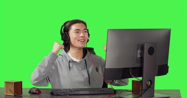 Asian person being on computer at desk, playing online rpg games with his friends. Young man having fun with new web contest, talking to people on headset over greenscreen backdrop. photo