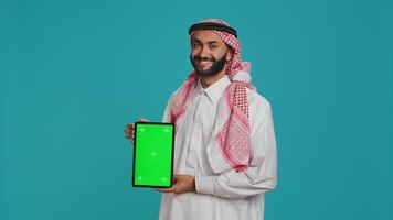Arab person shows greenscreen display over studio background, presenting isolated chromakey mockup template. Young guy in islamic attire holding tablet with blank copyspace screen. photo