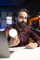 Smiling tech guru reviewing smart light bulb in studio, discussing with audience about home improvement. Cheerful man sharing latest news in WiFi home automation industry photo