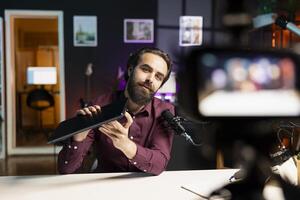 Man presenting laptop to audience, filming with professional camera and mic. Content creator assessing notebook product, recording with specialized equipment in studio photo