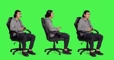 Asian person plays with controller while seated on chair against a full body greenscreen backdrop. Man engages in electronic gaming adventure with colleagues, gamer performs online games. photo