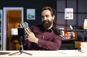 Tech expert presenting V mount battery, filming technology review using smartphone on tripod and professional microphone. Man showcasing charging device specs, recording with mobile phone photo