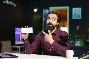 Man pointing at isolated screen cellphone in hand, using it as empty copy space to showcase his online streaming channel logo. Joyful media star does introductory using mockup phone photo