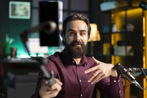 Content creator talking in front on cellphone attached to selfie stick in home studio interior, chatting with viewers. Man using phone camera to record vlog in rgb illuminated apartment photo
