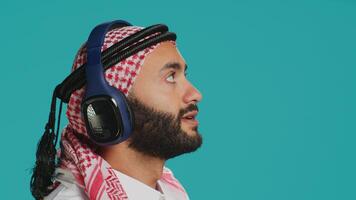 Cheerful guy dancing around in studio, listening to modern tunes on headphones and having fun on camera. Middle eastern adult in traditional clothes showing groovy dance moves and singing. photo