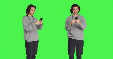 Guy texting messages on mobile phone standing against greenscreen backdrop, using social media app to chat remotely with friends. Asian joyful adult messaging on smartphone. photo