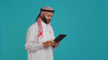 Young adult navigating on gadget in studio, browsing websites online on modern device. Muslim guy in traditional costume using tablet to check internet pages, chatting on social media. photo