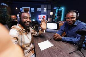 Interviewer waving towards cell phone and introduces guest, beginning a live talk show. Male influencers using mobile device and recording equipment for radio program and vlogging for online audience. photo