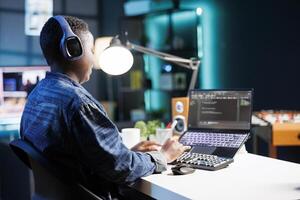Female freelancer listening to music while coding software and managing data. Black woman uses laptop to control and interact with network systems, ensuring secure communication. photo