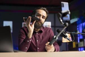 Tech expert assessing smartphone performance, talking about components used inside, filming with selfie stick. Entertainer unpacking mobile phone to review it on technology internet channel photo