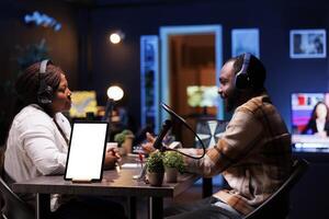Black man and woman with headphones making live broadcast while digital tablet displays blank white screen. African vloggers using podcast equipment while device placed on desk shows mockup template. photo