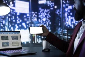 Company owner using smartphone showing white screen in office, sitting at his posh desk and admiring metropolis view. Financial advisor looks at blank mockup display on phone, business work. photo
