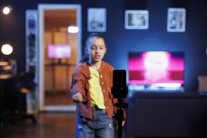 Focus shot on smartphone attached to tripod used by kid to do viral dance for social media viewers while listening to popular hit song. Girl in blurry background using phone to film dancing photo
