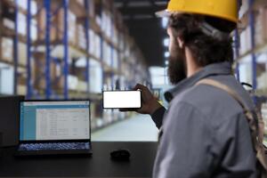 Warehouse worker presenting at phone screen with white layout, handling order receipts and invoices for distribution. Depot engineer managing cargo stock pallets for supply chain delivery. photo