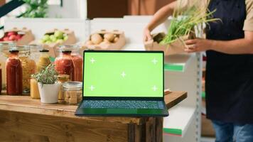 Laptop displaying greenscreen in bio shop filled with freshly harvested fruits and vegetables, sustainable lifestyle concept. Computer shows isolated chromakey template in zero waste store. photo