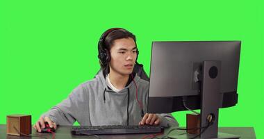 Young player sitting at computer desk and playing online games with friends, using modern devices to have a good time. Asian man participating in rpg tournament over greenscreen. photo