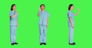 Medical assistant presents good sign over full body greenscreen backdrop, showing agreement and like gesture. Healthcare employee in uniform doing approval sign in studio. photo