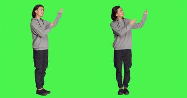 Asian man doing advertisement in studio, pointing at something next to him for new ad slogan. Young person smiling and working on promotional marketing, standing over full body greenscreen backdrop. photo