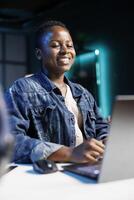 Close-up of African American lady smiling enthusiastically while typing on a digital laptop. Joyful black woman sitting at desk and using personal computer, researching and reviewing emails. photo
