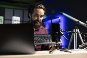 Influencer excited about newly released wireless keyboard, feeling impressed by high quality keys. Smiling man trying out office computer peripheral, filming using mobile phone on tripod photo