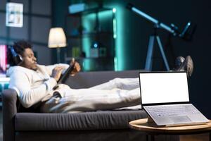 Man enjoying himself, searching new tv series to watch in apartment living room, focus on mockup laptop. Relaxed person wearing headphones, lying on couch next to isolated screen device photo