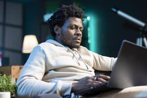 Self employed man working from home using laptop to send emails. African american man typing on notebook keyboard, composing documents in order to deliver finalized project to client photo