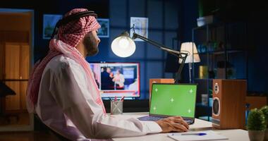 Middle Eastern man scrolling on green screen laptop, checking social media posts. Self employed man browsing online websites from mockup device in apartment office with TV as background noise photo
