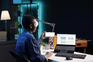Female freelancer holding smartphone with white screen and using laptop. Black woman sitting in front of her personal computer while horizontally grasping cell phone with blank chromakey template. photo
